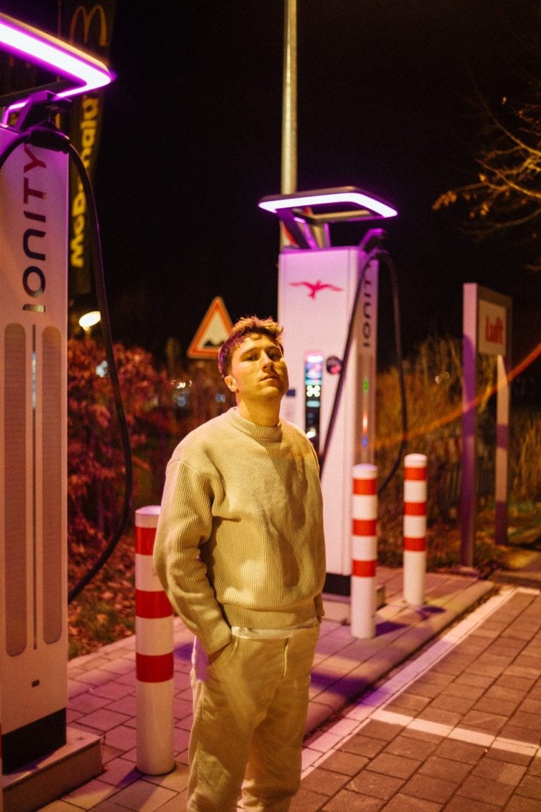 Ein junger Mann in beigem Pullover steht nachts vor beleuchteten Ionity Ladestationen auf einem Parkplatz mit rot-weißen Absperrpfosten.