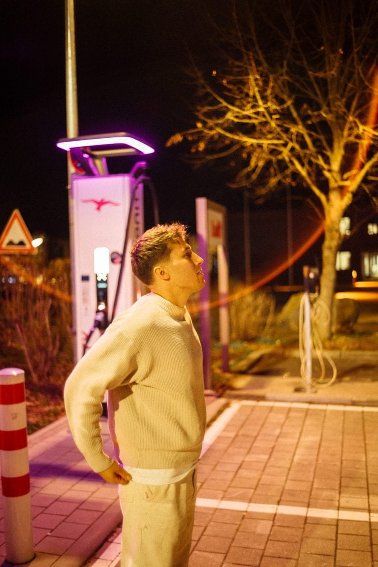 Ein junger Mann in einem beigen Pullover steht nachts nachdenklich vor einer beleuchteten E-Ladestation auf einem gepflasterten Parkplatz.