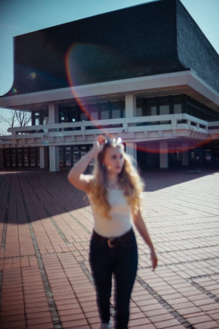 Eine junge Frau mit langen blonden Haaren läuft unscharf auf einem gepflasterten Platz vor einem modernen, architektonisch markanten Gebäude.