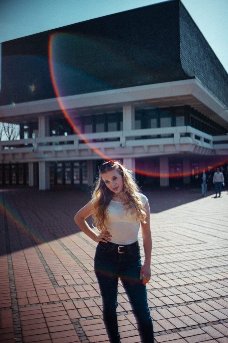 Eine junge Frau mit langen blonden Haaren posiert vor einem modernen, architektonisch markanten Gebäude bei hellem Sonnenlicht mit Lens Flare.
