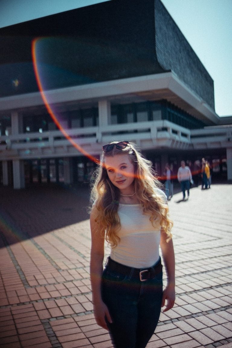 Eine junge Frau mit langen blonden Haaren und Sonnenbrille steht vor einem modernen Betongebäude bei sonnigem Wetter mit einem markanten Lichteffekt.