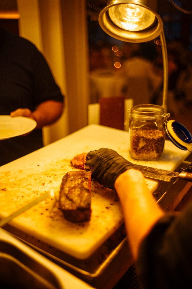 Eine Person mit schwarzen Handschuhen schneidet ein gegrilltes Steak auf einem weißen Schneidebrett unter einer warmen Wärmelampe in einer Küche.