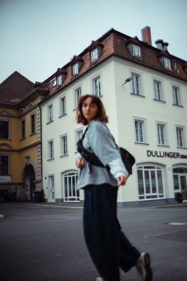 Eine junge Frau mit braunen Haaren läuft unscharf im Vordergrund vor einem hellen, historischen Gebäude mit vielen Fenstern in einer Stadt.