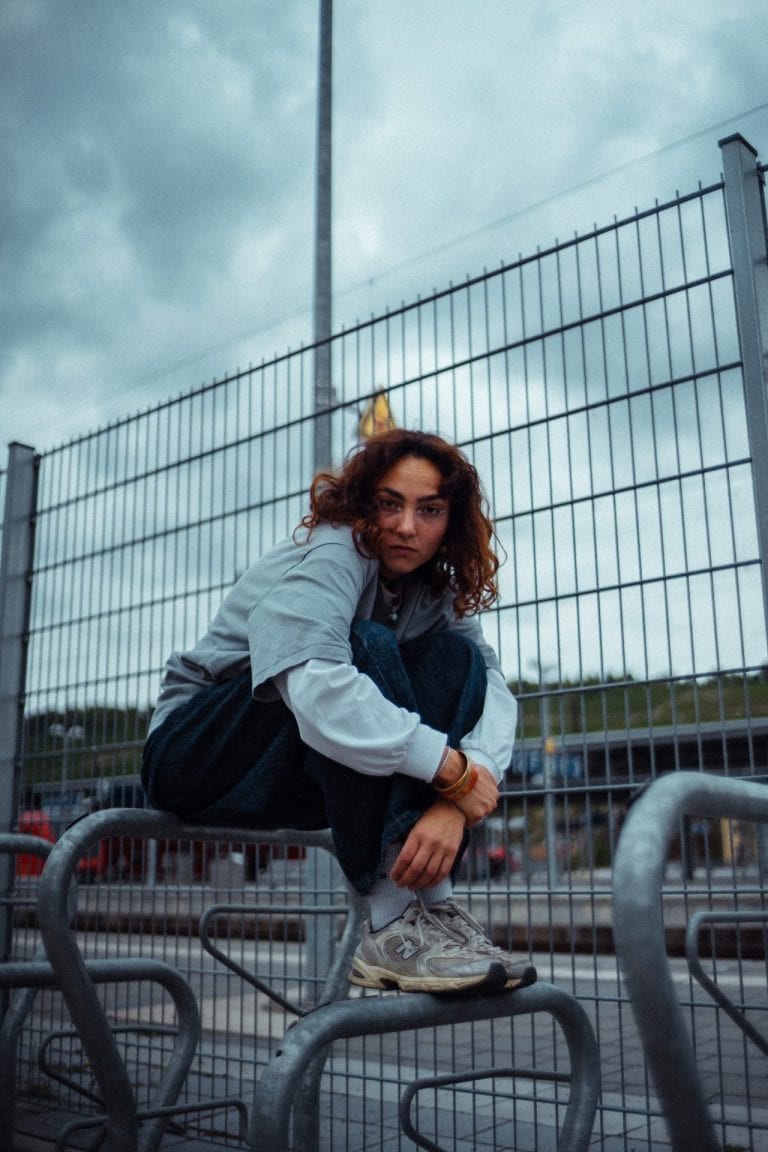 Eine junge Frau mit lockigem Haar sitzt auf einem Fahrradständer vor einem hohen Metallzaun unter einem bewölkten Himmel in urbaner Umgebung.