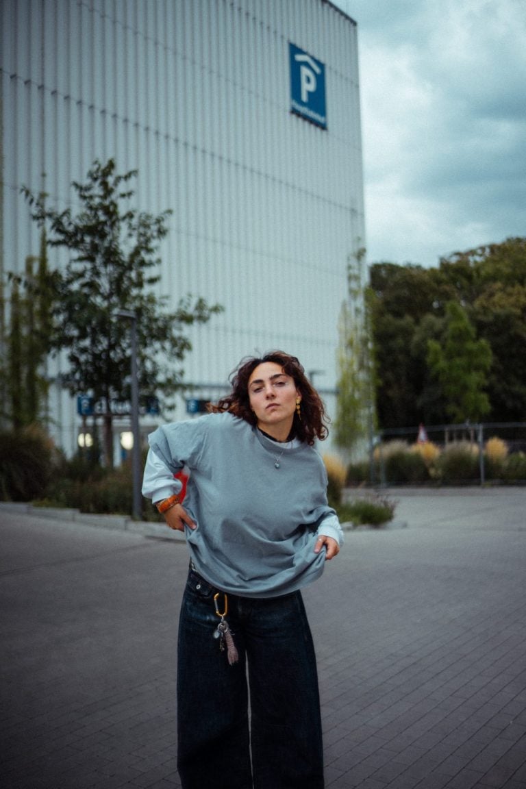 Eine junge Frau mit lockigem Haar trägt ein graues Sweatshirt und eine weite Jeans vor einem modernen Parkhaus mit blauem P-Schild im Hintergrund.