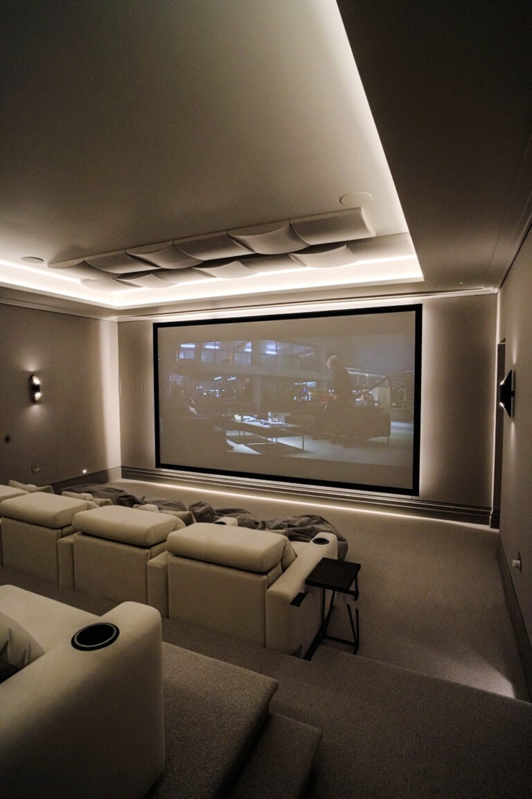 A modern home theatre room with tiered seating, white sofas, and a large screen displaying a film scene, illuminated by soft recessed lighting—an example of luxury home cinema installation in Roehampton Gate Surrey.