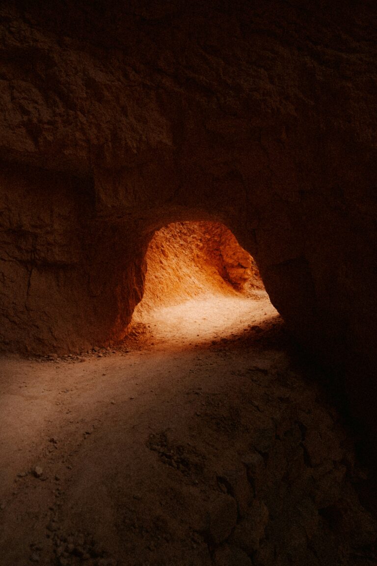 Narrow rocky tunnel with an illuminated dirt path and reddish-brown walls.