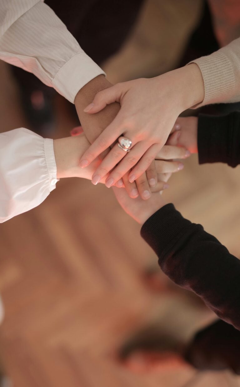 A group of hands stacked together in a show of teamwork, with different colored sleeves and one hand wearing a ring.