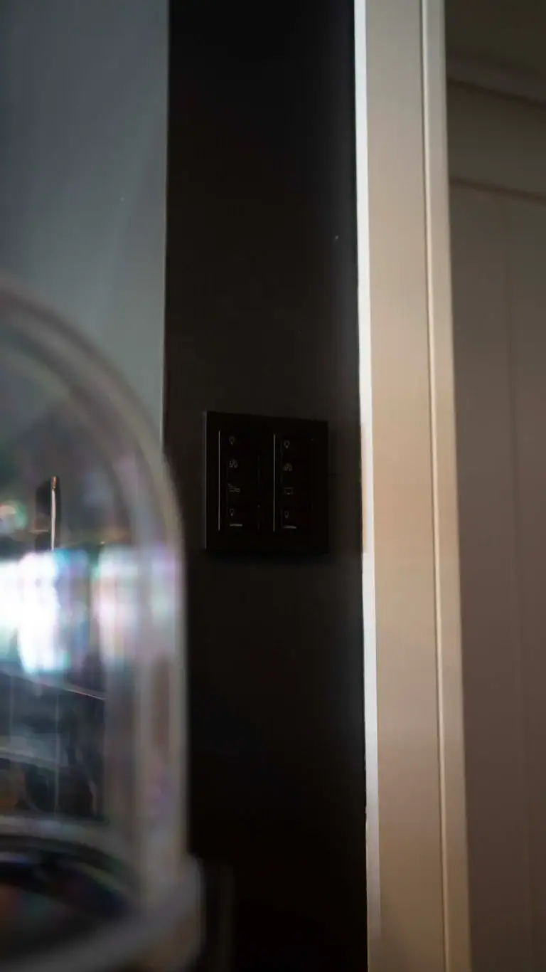 A black light switch panel, part of smart home automation in Epsom Surrey, is mounted on a dark wall next to a white door frame; a blurry, transparent object appears in the foreground on the left side.