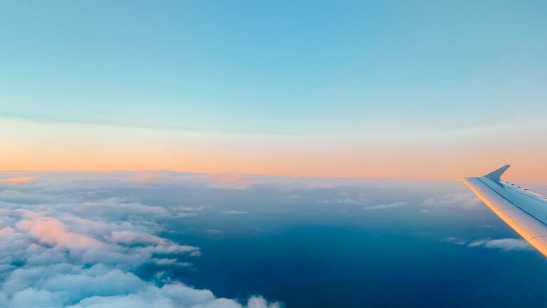 View from an airplane window showing a wing above soft clouds at sunrise, representing the experience of flying with a disability and accessible air travel. Featured image for the blog post "Flying with a Disability".