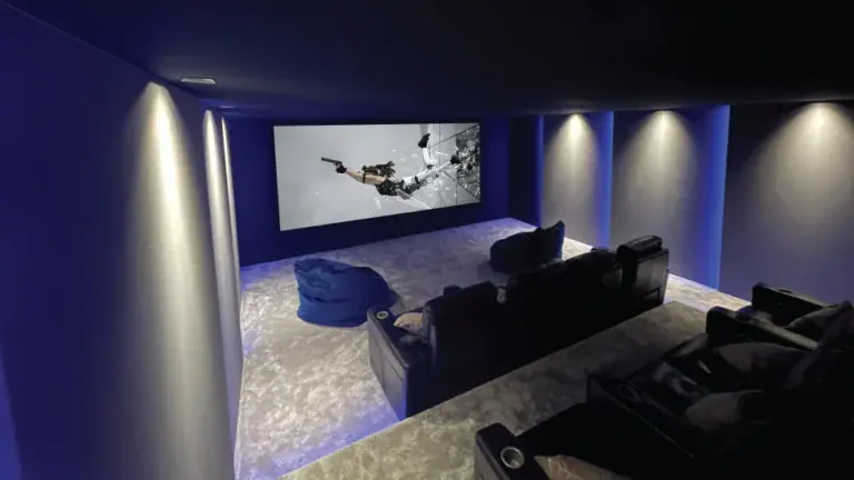 Experience a modern home theatre room with dark walls, recliner seats, a large screen showcasing an action film, soft carpet flooring, and ambient blue lighting—an ideal home cinema sample in our Surrey showroom.