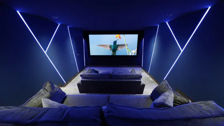 A modern home theatre room—perfect for a premium home cinema installation in Guildford, Surrey—with blue LED lighting, plush grey sofas, and a large screen displaying a close-up of a hummingbird in flight.