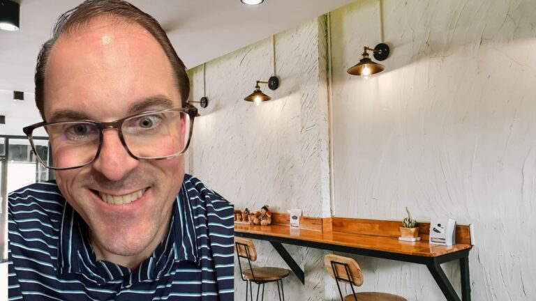 Close-up of Travis with glasses in a modern indoor setting, featuring a textured wall, wooden counter, light fixtures, and small decor items.