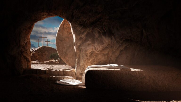 Interior of a cave with a stone slab and cloth, looking out to three crosses on a hill.