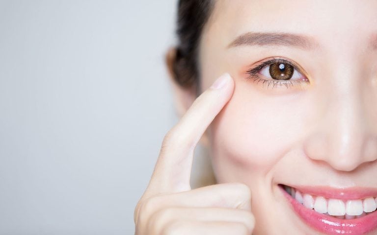 Smiling woman pointing to her eye area, highlighting smooth skin and defined eyelid crease after blepharoplasty