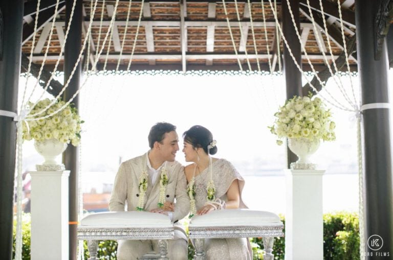 Sirikarn and Nicholas in Thai outfits laugh under a white floral pavilion at Anantara Riverside Bangkok Resort