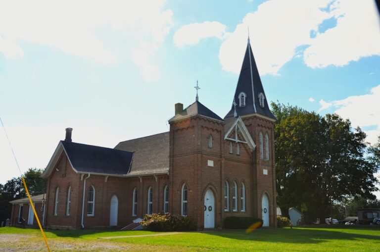 A brick church with a tall steeple and arched windows, set against a blue sky with clouds.