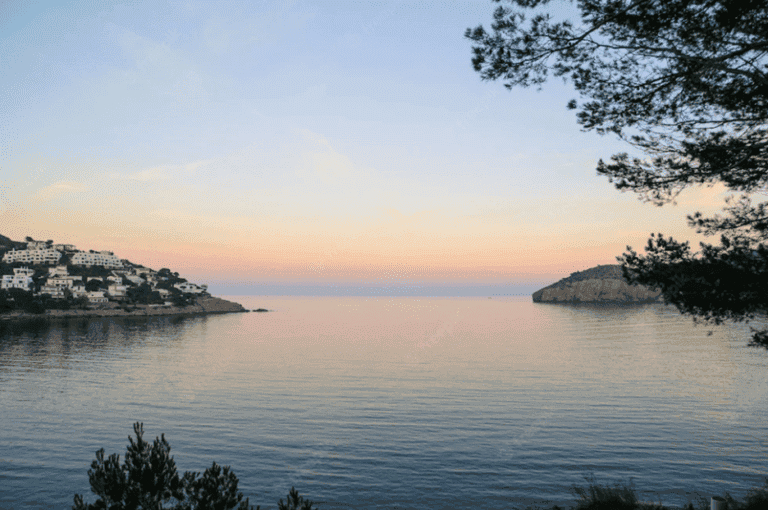 Serene coastal landscape at sunset with calm waters, lush trees, and charming hillside villas, perfect for relaxing vacations and scenic getaways in Can Camades, Mallorca.