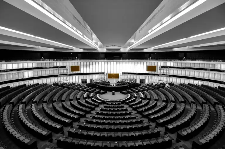 A black and white image of a government hall with lots of chairs. Three orange tv's are hanging on the wall