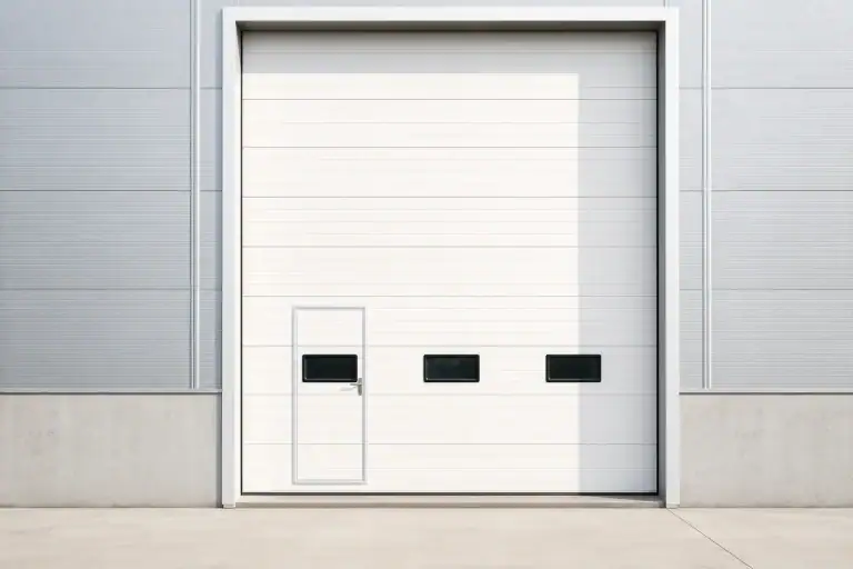 White custom commercial sectional door featuring an integrated wicket door for pedestrian access and rectangular vision windows, installed on a modern factory facade.
