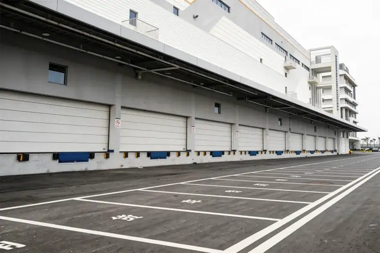 Exterior view of a massive modern logistics center featuring a continuous row of heavy-duty industrial sectional doors and blue dock levelers, demonstrating the high-volume manufacturing and delivery capacity required for mega projects.