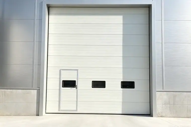 White commercial sectional overhead door featuring built-in windows and a pedestrian wicket door installed on a modern industrial building exterior.