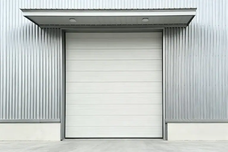 White commercial sectional overhead door installed on a modern industrial warehouse with corrugated metal siding and an overhead canopy.