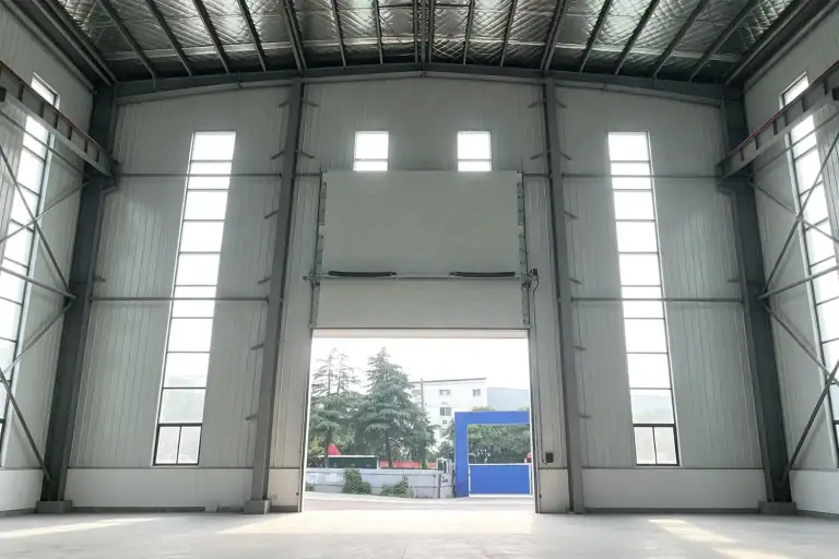 Interior view of a modern industrial warehouse featuring steel framing, high vertical windows, and a partially open sectional overhead door.
