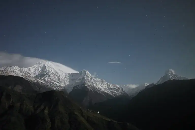 Snow-capped mountains under a starry night sky, with dark forested hills in the foreground and a soft layer of clouds partially covering the peaks.