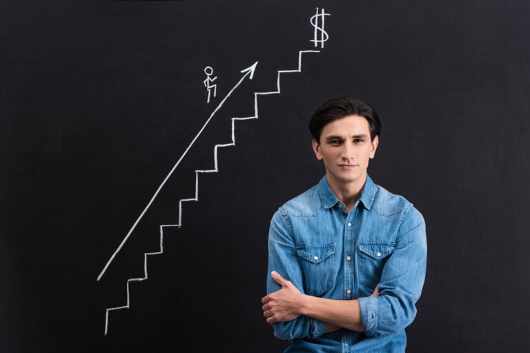 Man wearing a denim shirt standing in front of a blackboard that has a white chalk illustration of steps leading to a $. An arrow with a stick figure man points up the steps.
