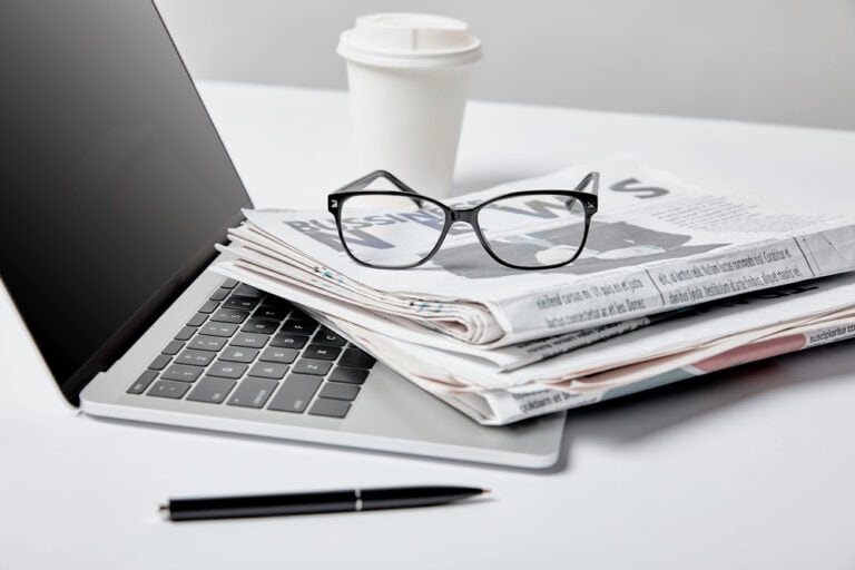 Selective focus of laptop with blank screen near business newspapers, glasses, pen and paper cup on white"