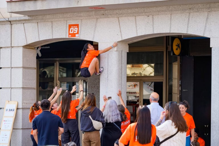 Una escaladora en camiseta naranja sube por un pilar mientras un grupo la anima en el centro de Torrejón de Ardoz.