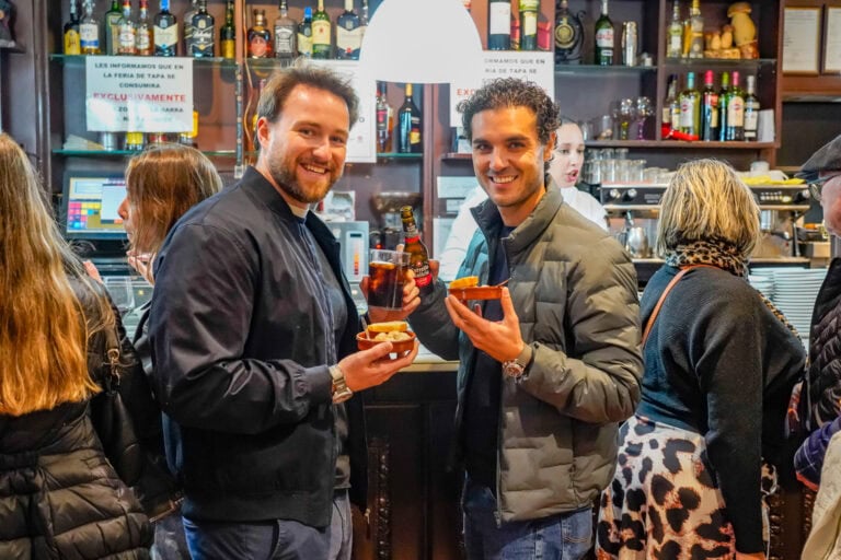 Dos hombres sonrientes sostienen tapas y bebidas en un bar, mientras otros clientes están de fondo. Contexto en Torrejón de A