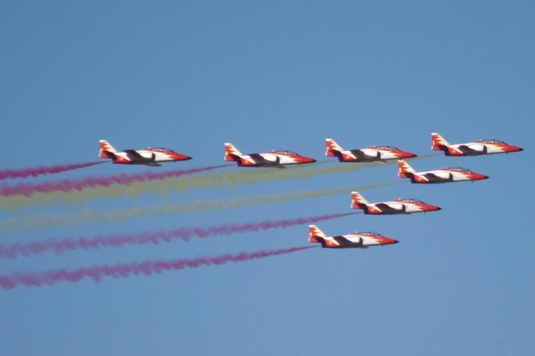 Un grupo de aviones en formación vuela en el cielo, dejando estelas de humo de diferentes colores. La imagen capta la acrobac