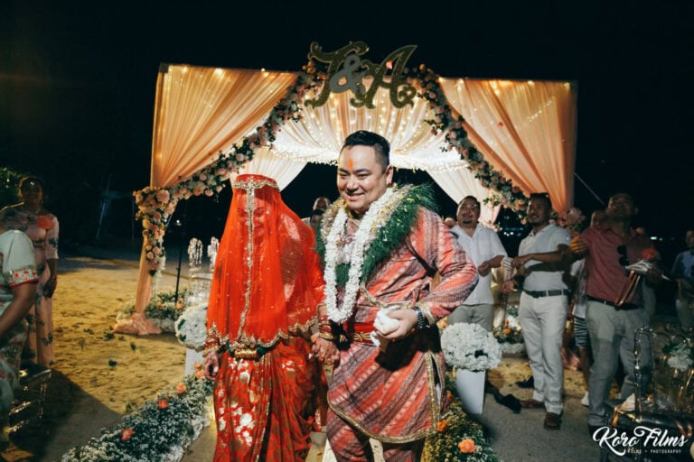 The veiled bride and the groom in garlands stand in front of the mandap at night in Pattaya
