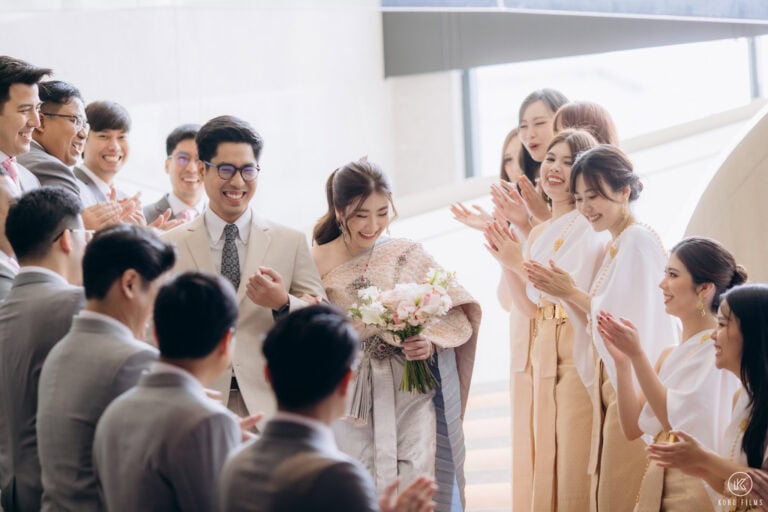 Pla and Patt walk in as bridesmaids in Thai dress clap at their Thai engagement ceremony at Carlton Hotel Bangkok Sukhumvit