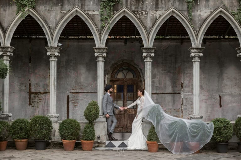 Nagaya’s long veil flies as she and Amarvir hold hands in front of gothic stone arches at Benedict Studio, Bangkok