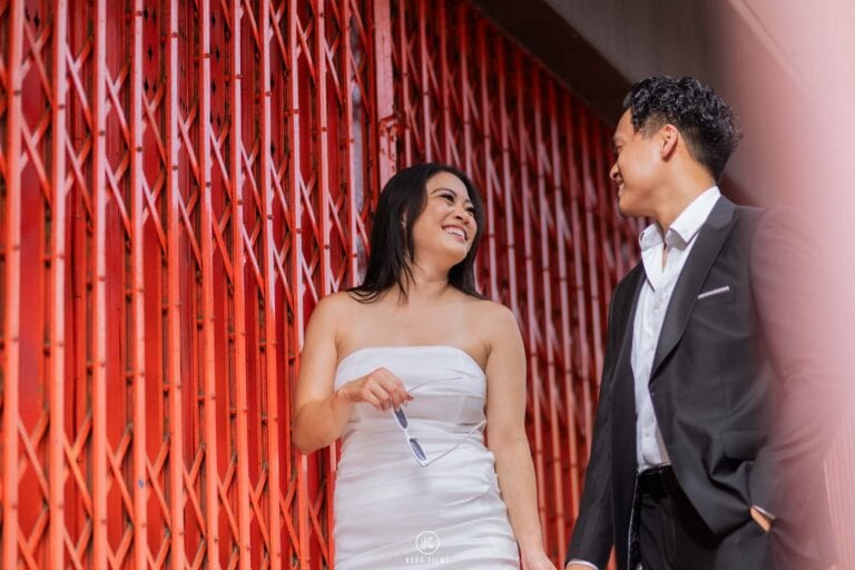 Tracy in a white strapless dress and Lan in a dark suit smile in front of red folding shutters in Chinatown, Bangkok