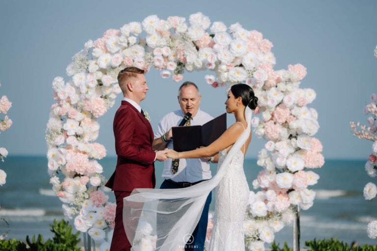 Jakub and Clear say their vows with the celebrant under a floral ring arch on the seafront at Sheraton Hua Hin
