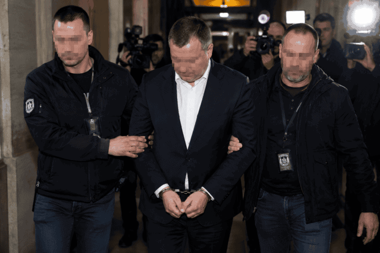 Bilincsben vezetnek egy öltönyös politikust egy hivatali épület folyosóján, újságírók és kamerák előtt.