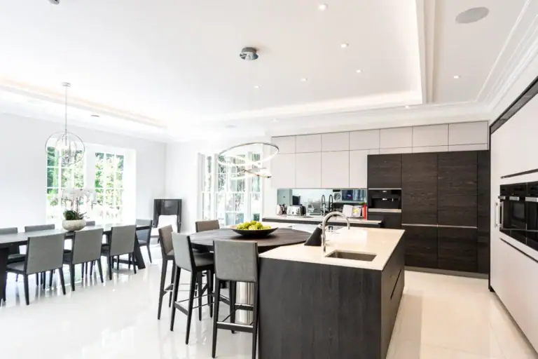Modern kitchen with a large island, dark cabinetry, and built-in appliances features home automation with smart lighting and shading installation. A dining table with grey chairs sits by large windows, filling the space in Cobham, Surrey with natural light.