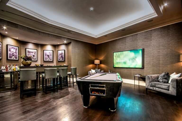 A modern basement home cinema with bar in Weybridge, Surrey, featuring a pool table, sleek bar area with stools, framed photos, a large TV, grey sofa, and dark wooden flooring under recessed lighting.