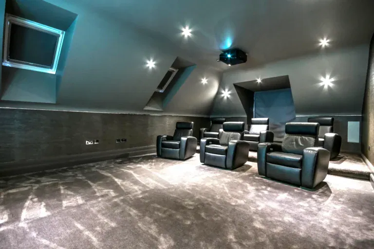 Modern home theatre room with grey walls and carpet, featuring six black leather reclining chairs in two rows, a ceiling-mounted projector, and soft recessed ceiling lights—perfect for a luxury home cinema installation in Oxshott.