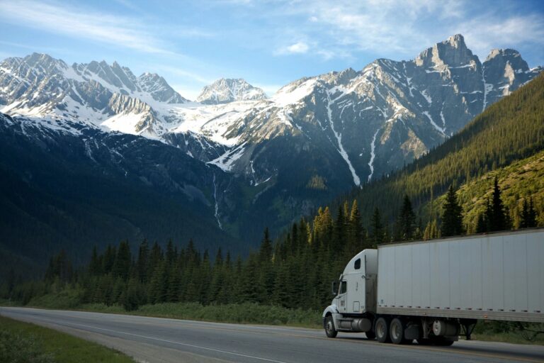A white semi truck driving on a highway with snow-capped mountains in the background, representing reliable trucking and logistics support services.