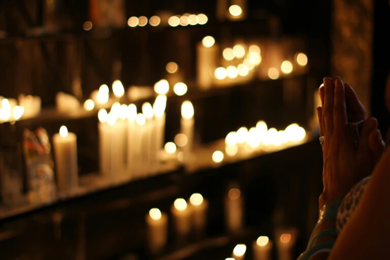 A warmly lit scene with numerous lit candles on shelves, creating a soft glow. Hands are clasped in prayer, evoking a calm and reverent atmosphere.