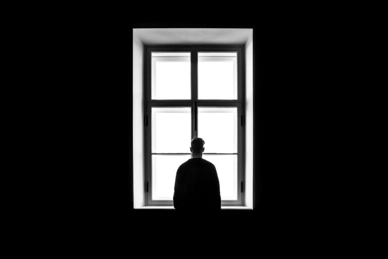 A silhouette of a person standing in front of a bright, four-pane window in a dark room.