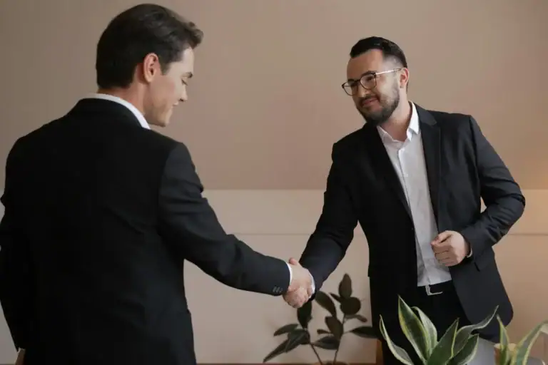 Two business professionals in suits shaking hands and smiling at each other.