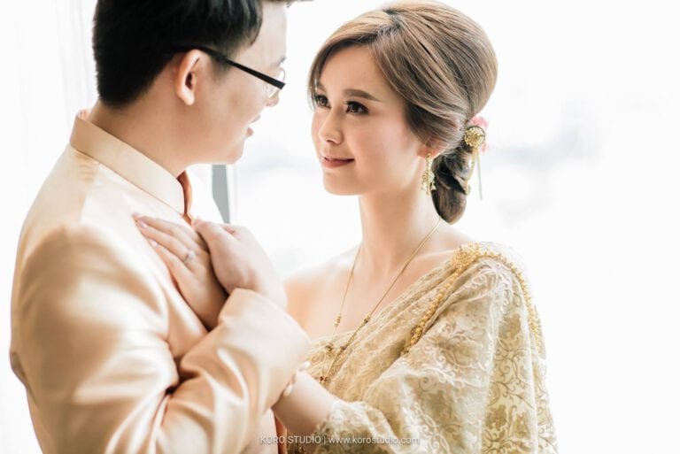 Lily and Darren in gold Thai wedding outfits embrace by a window at InterContinental Bangkok