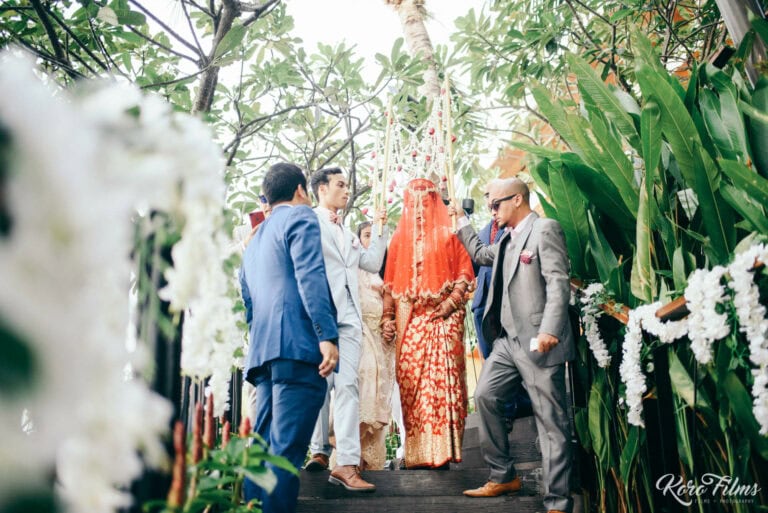 Family members walk the veiled bride in red through white orchids to the ceremony in Pattaya