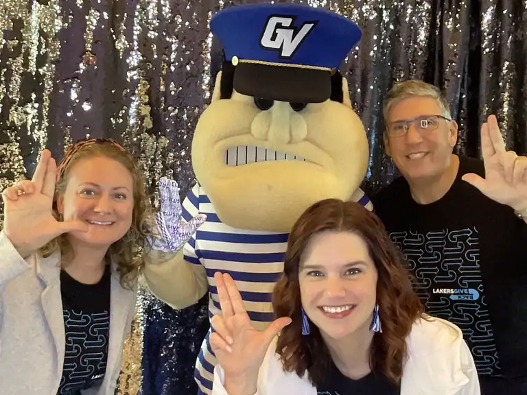 Mascot in blue striped shirt and GV cap posing with three guests wearing “LAKERS GIVE” shirts at Fastbooth backdrop in Grand Rapids, MI.